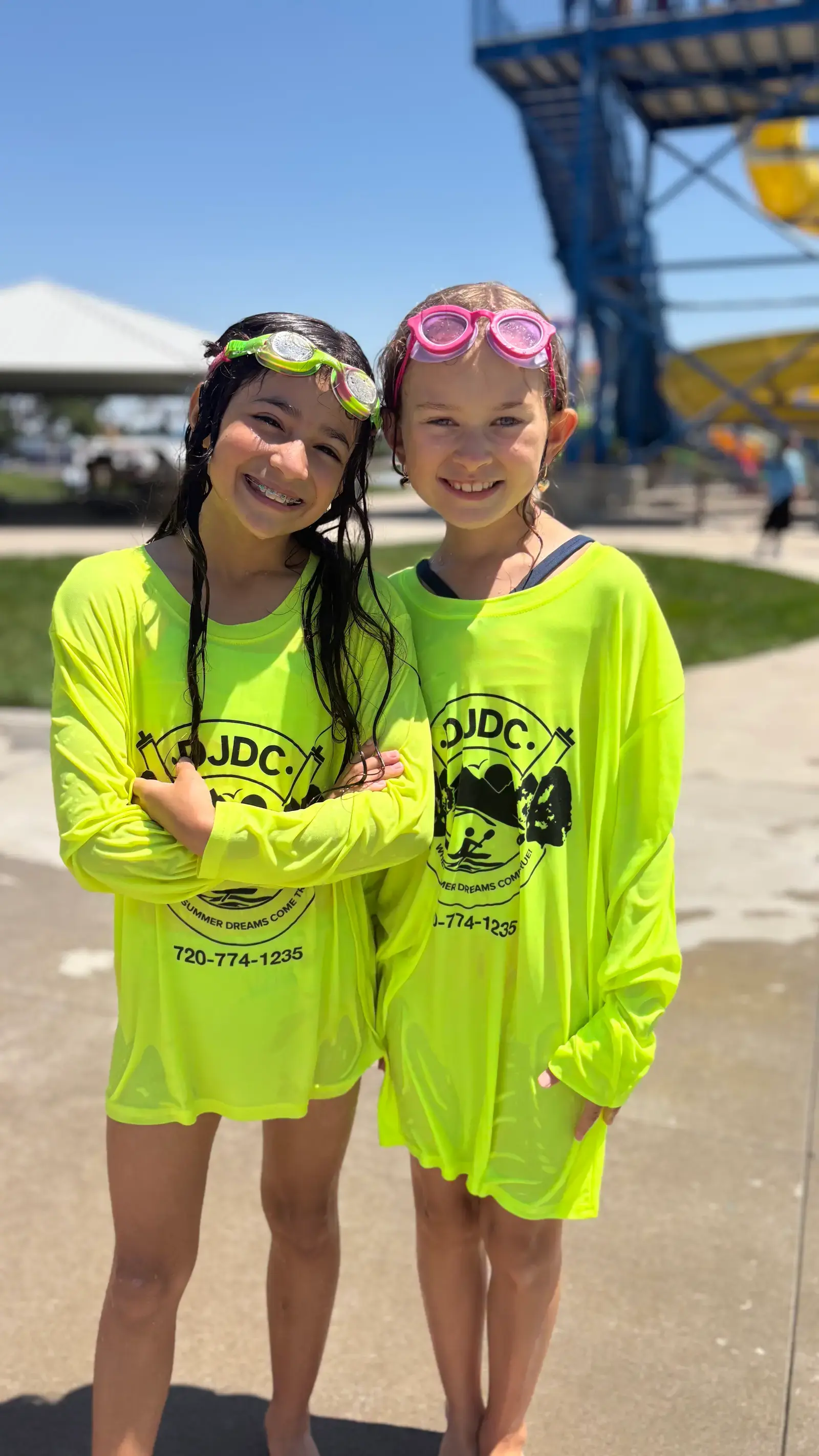 Two campers in neon camp shirts with swim goggles pushed up on their heads, grinning