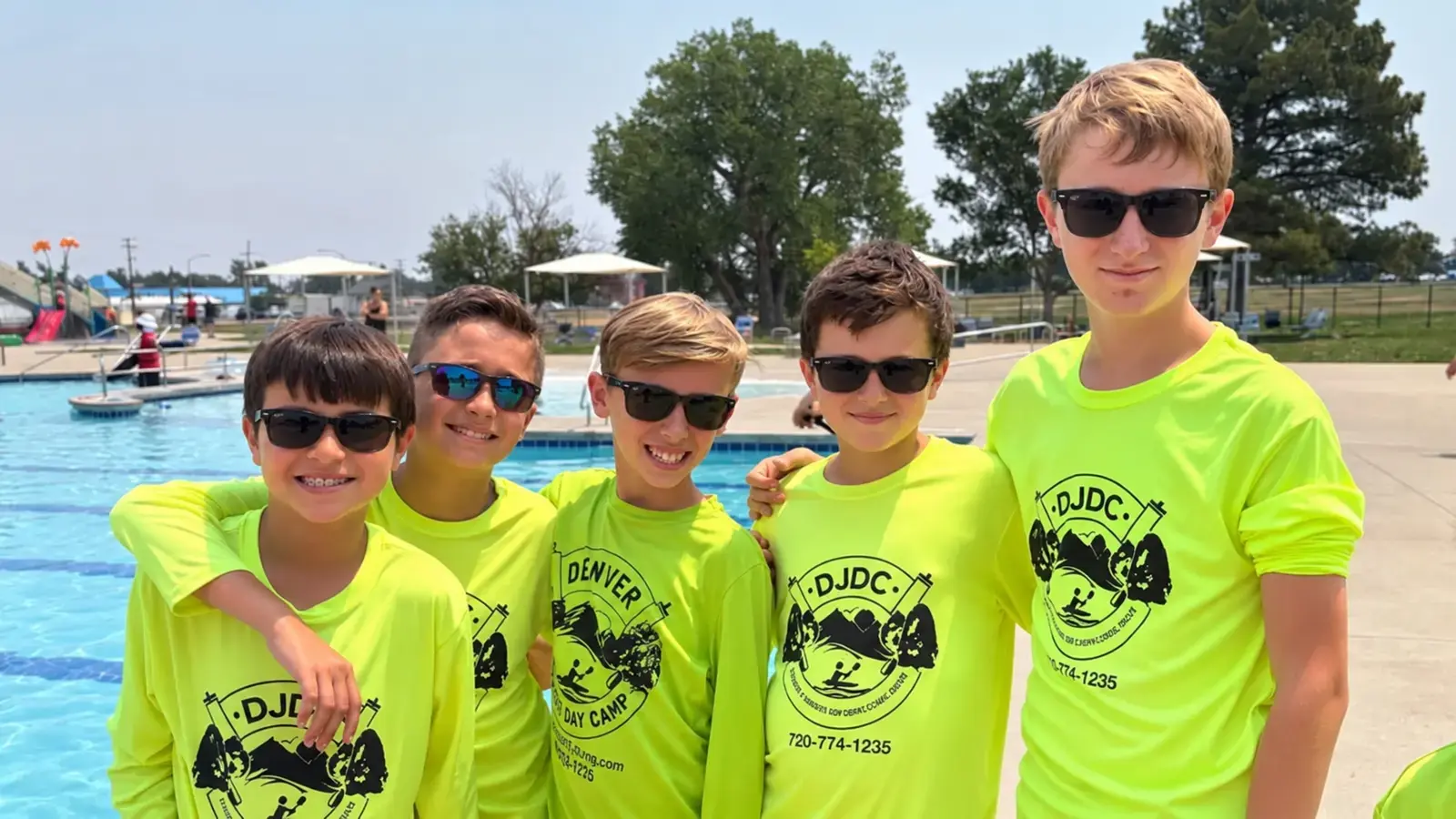 Five campers in long-sleeve DJDC rash guards and sunglasses beside the pool