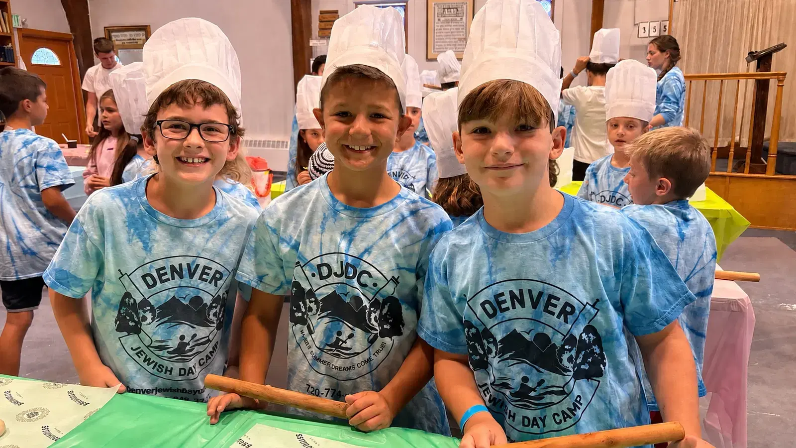 Three campers in chef hats and tie-dye camp shirts holding rolling pins at the baking table