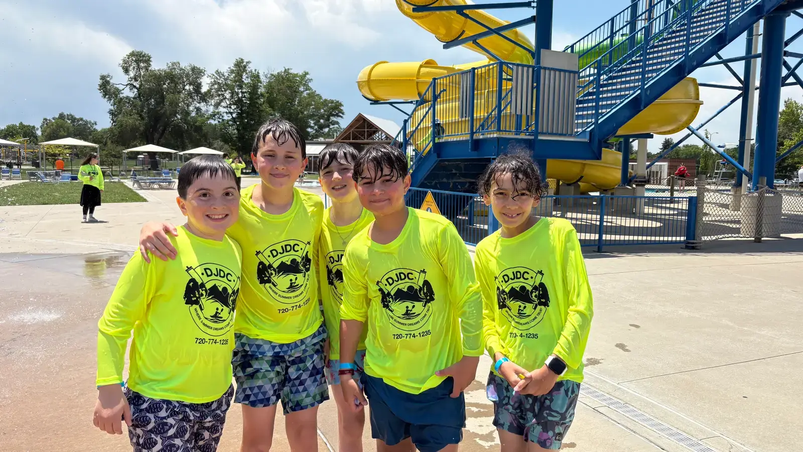 Five campers in neon DJDC shirts with arms around each other in front of the water-park slide