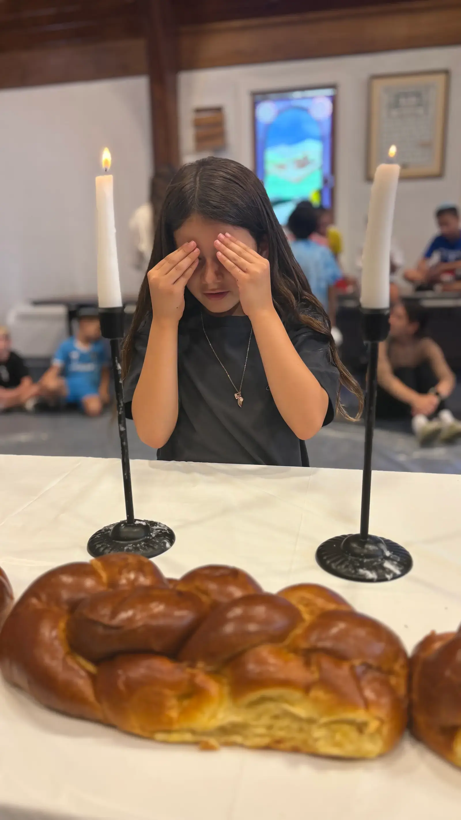 A camper covering her eyes for the blessing over the Shabbat candles, fresh challah on the table