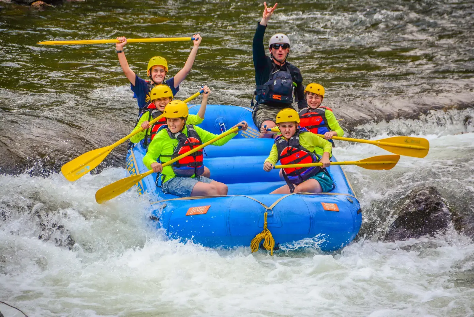Campers in helmets and life jackets cheering with paddles raised as their raft runs the whitewater